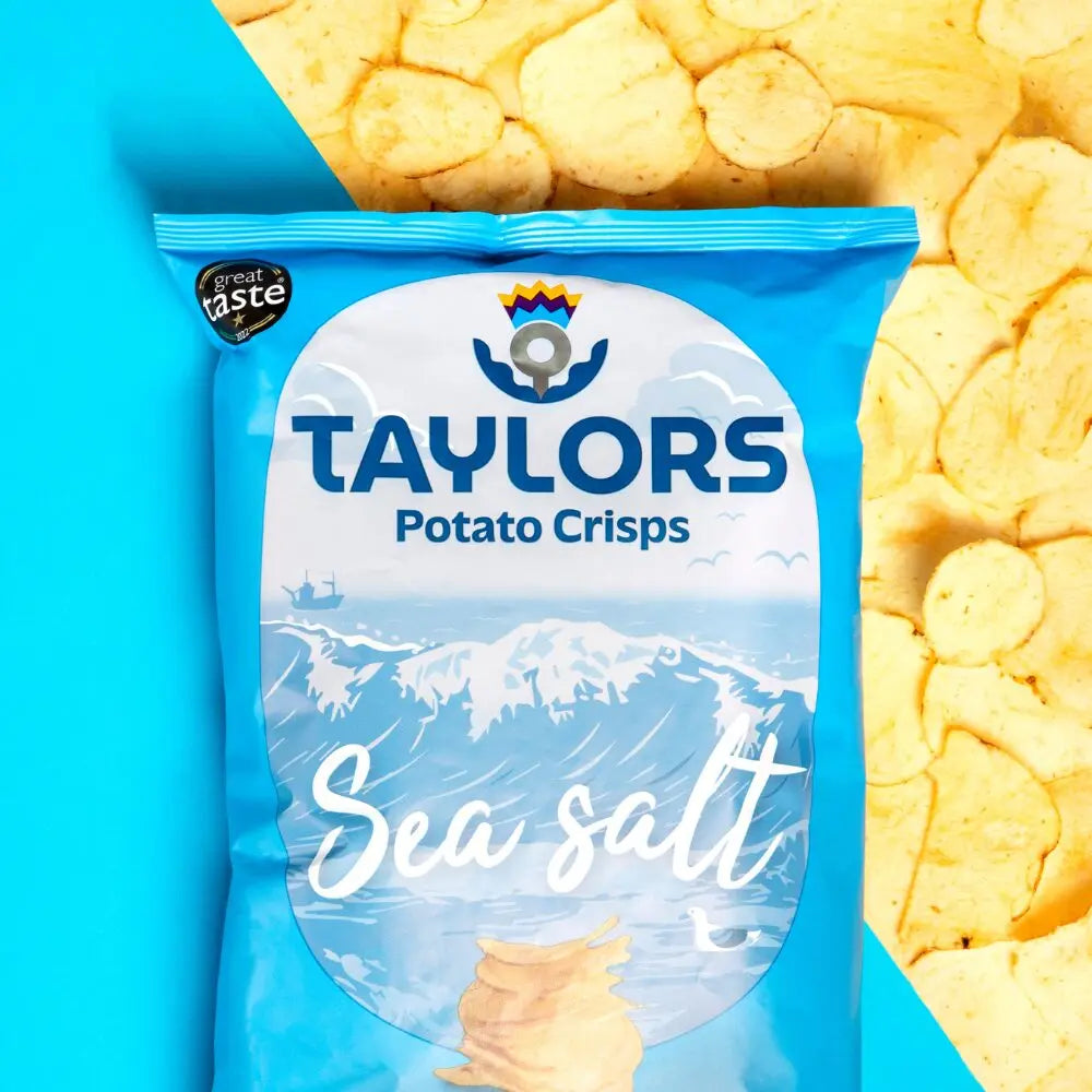 A bag of Taylors Potato Crisps Sea salt on a bright blue background with a close-up of potato crisps in the top right corner. The bag is light blue with a large white oval label featuring the brand name and a wave graphic.